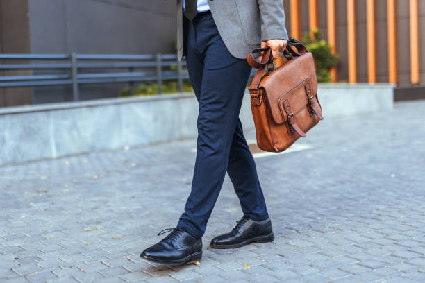A man in a business suit walks confidently on a city street carrying a brown leather briefcase. The focus is on his stylish formal shoes and elegant leather accessory, symbolizing professionalism.